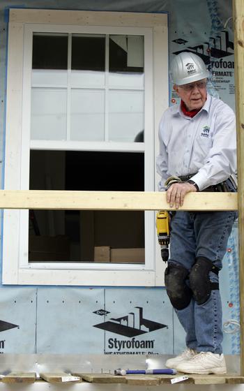 Jimmy Carter with kneepads on and with a drill in his hand stands in front of a home under construction.