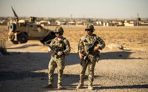 Two American soldiers armed with guns stand in a dry, desert climate in front of an armored military vehicle.