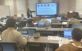 An instructor points at coursework on a large screen at the front of a room of military service members seated at tables with laptop computers.