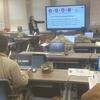 An instructor points at coursework on a large screen at the front of a room of military service members seated at tables with laptop computers.