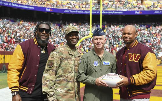 Four men stand on a football field, two on either end wearing official Washington Commanders football jackets, and the two in the middle wearing Air Force uniforms, together hold a white football with red lettering.