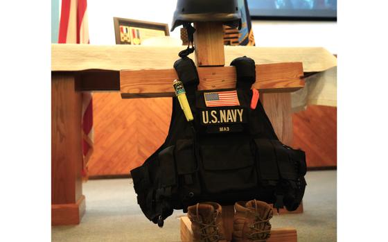 A duty vest with “U.S. Navy” and a U.S. flag, boots and a helmet adorn a wooden cross.