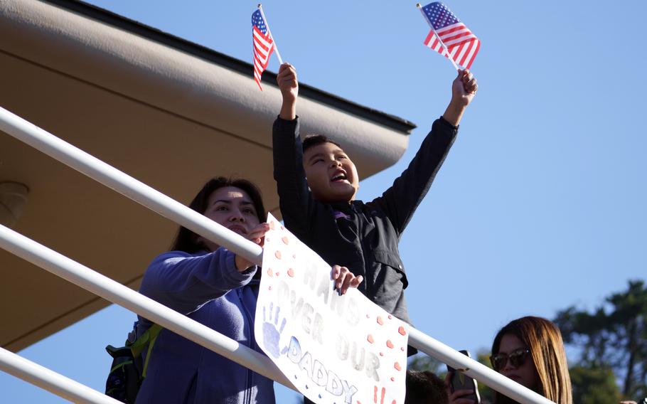 On a pier, a small boy raises two small US flags in his hand while a woman crouches next to him holding a sign reading, “Hand Over Our Daddy.”