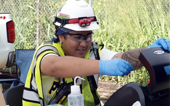 A woman in a yellow reflective vest, white hard hat and blue rubber gloves takes a water sample from a well.