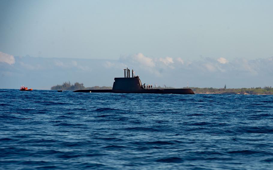 The top of a submarine is seen above the water surface line with coastal land in the background.