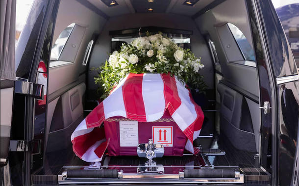 Flowers and the American flag draped over a red casket in the back of a black van.