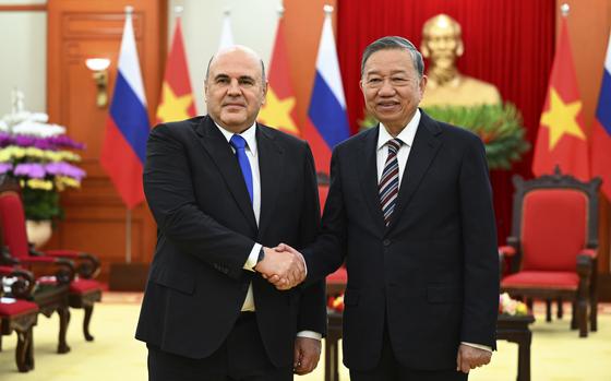 Two men in suits shake hands while facing the camera and smiling.