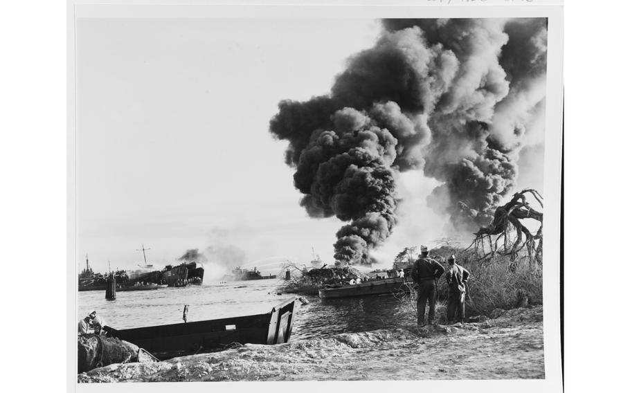 Men watch as USS LST-39 is afire in Pearl Harbor, Hawaii, after the May 21, 1944, explosion that destroyed several LSTs in the West Loch. Men are visible on her bow ramp. Other vessels are alongside and nearby engaged in firefighting. 