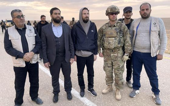 An American man in a hooded jacket stands next to a U.S. soldier in combat gear and Syrian officials on a road through a desert.