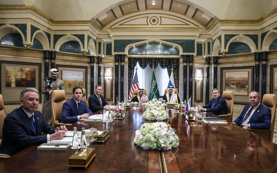 High-profile officials on a long table in a fancy room, with the U.S., Saudi Arabian and Russian flags lined up in the background.