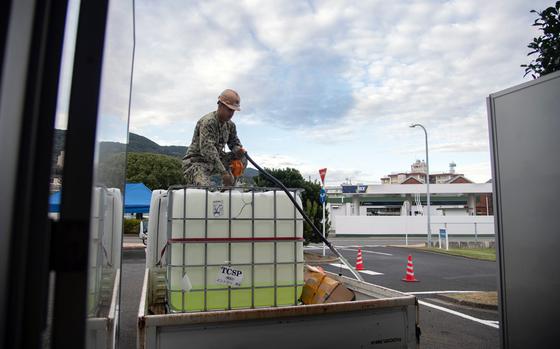 A man in a military uniform and hard hat kneels on top of a gas container in the back of a truck.