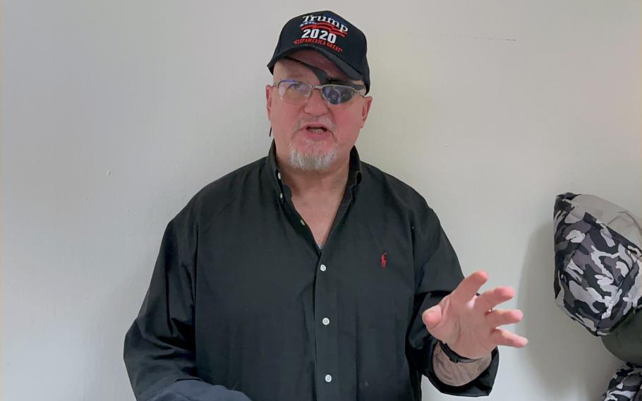 A man with an eye patch and a Trump hat talks against a white wall with one hand raised.