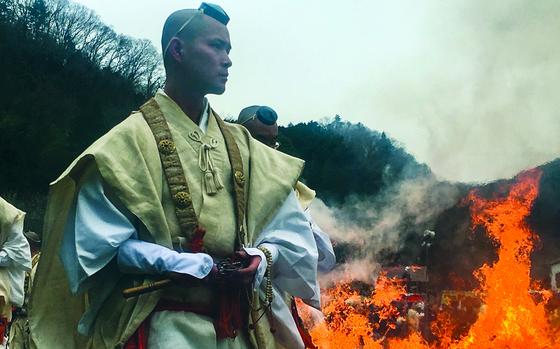 A Japanese monk in traditional attire stands in the foreground while a fire burns behind him.