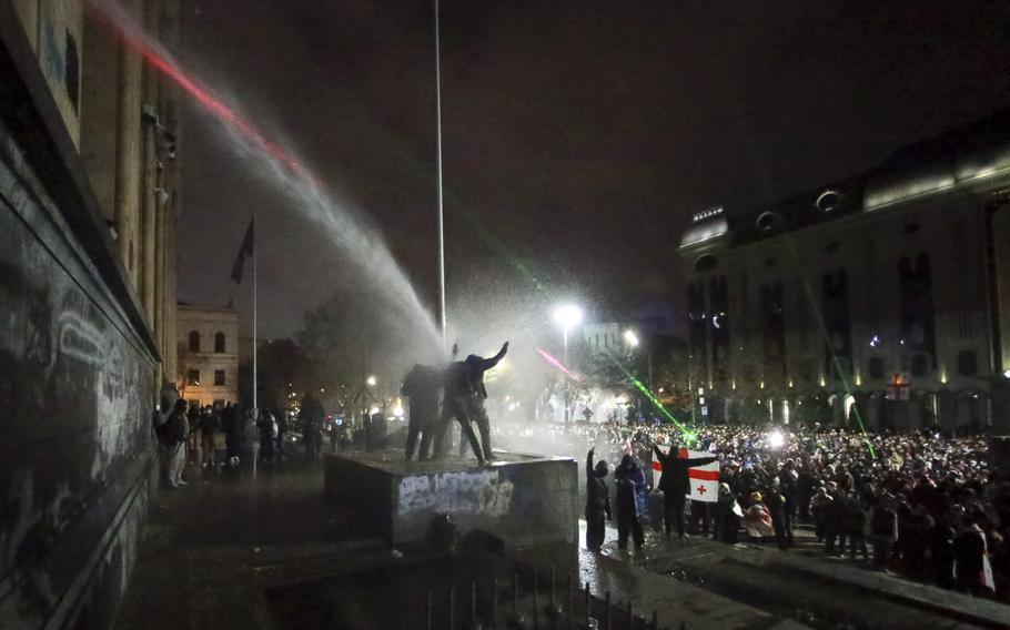 Protesters rally in Tbilisi, Georgia, at night, including three standing atop a tomb being sprayed by a water cannon.