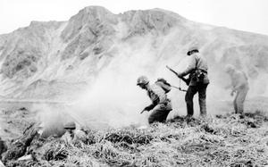 Army soldiers seen in black and white are carrying guns and walking down a grassy hill surrounded by gun smoke.