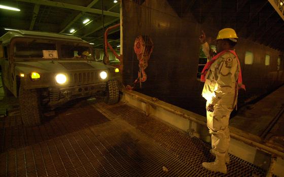 A soldier in a camouflage uniform, orange reflective vest and hard hat directs a Humvee on a ramp to a ship.