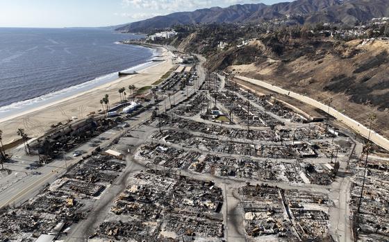 An aerial view of buildings destroyed by wildfires next to a ocean coastline with mountains in the background.