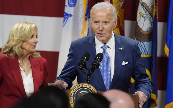 Joe Biden stands at a podium next to Jill Biden and speaks into microphones in front of U.S. and military flags.