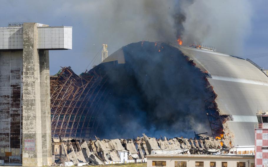 A large section of a blimp hangar is burned away as the structure smolders with fire.