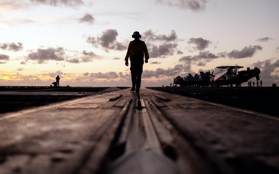 A sailor is silhouetted against the setting sun while walking along the flight deck of an aircraft carrier.