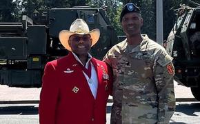 An Army soldier in camouflage uniform and black beret stands next to a man in a red blazer and cowboy hat.