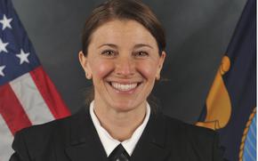 A Navy officer in dress uniform sits for a portrait in front of U.S. and Navy flags.