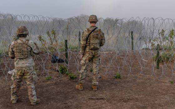 Two soldiers in camouflage uniforms and helmets, seen from behind, look through a barbed wire fence across an open grassy area.