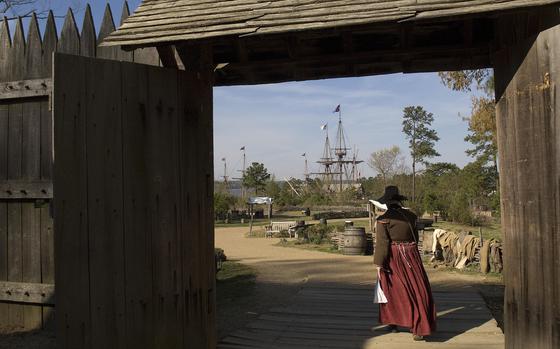 A woman in historical pilgrim garb walks through a rustic wooden gate.