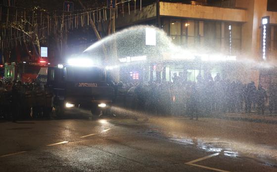 Police in riot gear stand in a line next to a large vehicle spraying a water cannon at protestors.