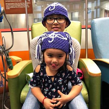 Two girls wearing purple knitted hats pose on a chair in a hospital.