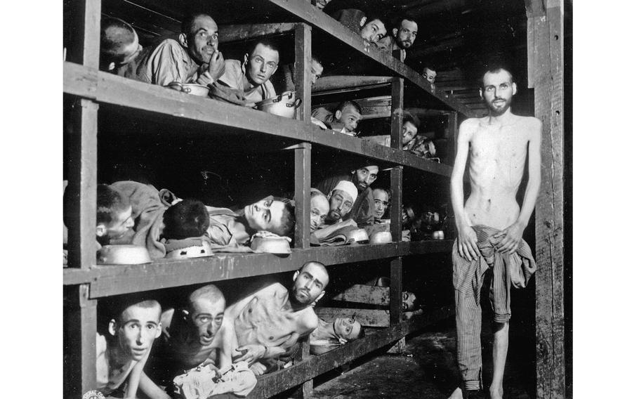 A black-and-white photo shows malnourished Holocaust survivors at KZ Buchenwald peering out of their four-level bunks after the liberation of the concentration camp. One man stands shirtless near the bunks.