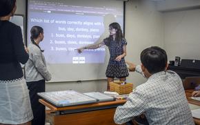 A teenage girl points to English text projected on a screen as a class member points at the screen and other students look on.