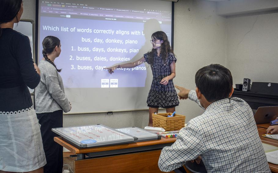 A teenage girl points to English text projected on a screen as a class member points at the screen and other students look on.