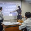 A teenage girl points to English text projected on a screen as a class member points at the screen and other students look on.