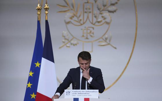 French President Emmanuel Macron speaks with one hand on his face and another on the podium in front of him. Two flags are behind him on the photo’s left.