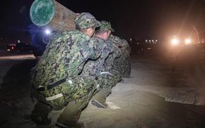 Navy SEALs crouch on a beach at night near the surf while carrying a log. 