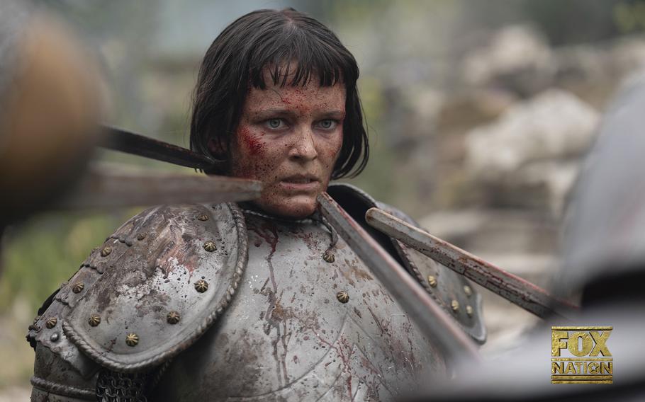 Joan of Arc, played by Liah O’Prey, her face smeared in blood, is dressed in armor, which is covered in dirt. She seems frightened by the long objects pointed at her face.  