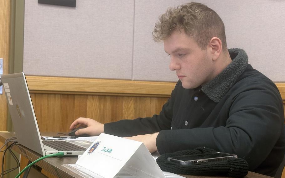 A man in a black jacket sits at a table and works on a laptop computer.