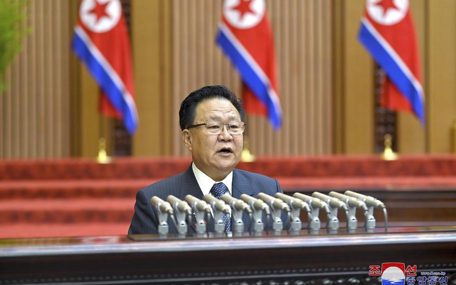 A North Korean official speaks in front of many microphones and three North Korean flags during an assembly.