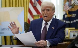 Donald Trump holds a document and gestures with his hand while seated at a desk in the Oval Office.