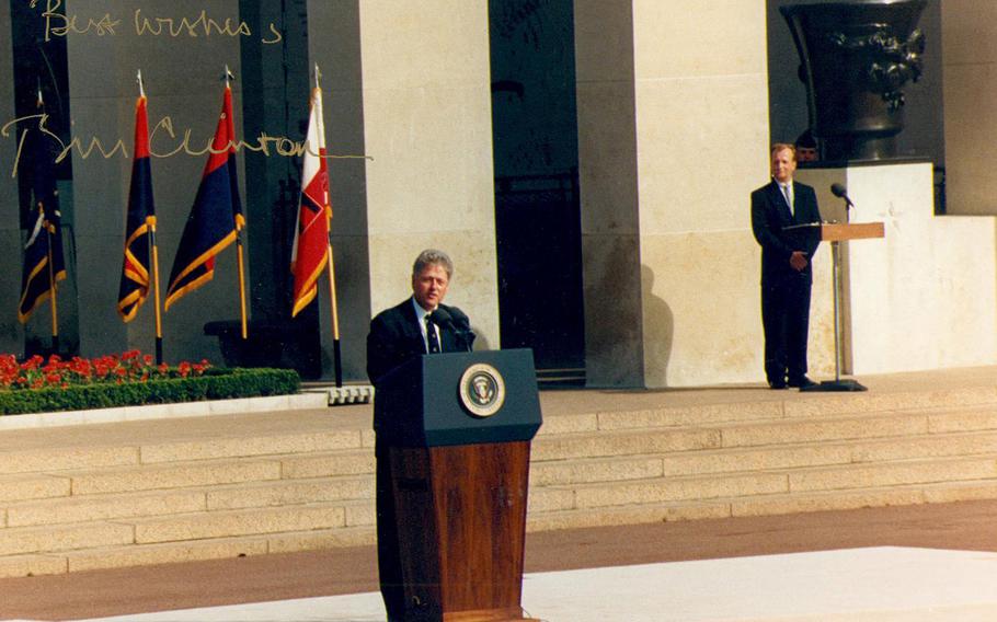 Former President Bill Clinton stands at a pedestal in the foreground while at right, in the background,  David Stewart looks on.