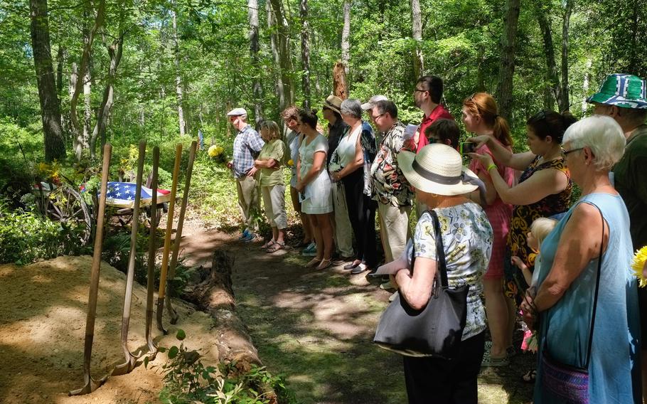 A line of mourners stand before a green burial gravesite in a forest.