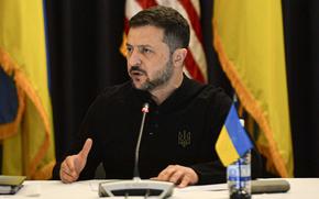 Volodymyr Zelenskyy sits at a table and speaks into a microphone in front of U.S. and Ukrainian flags.