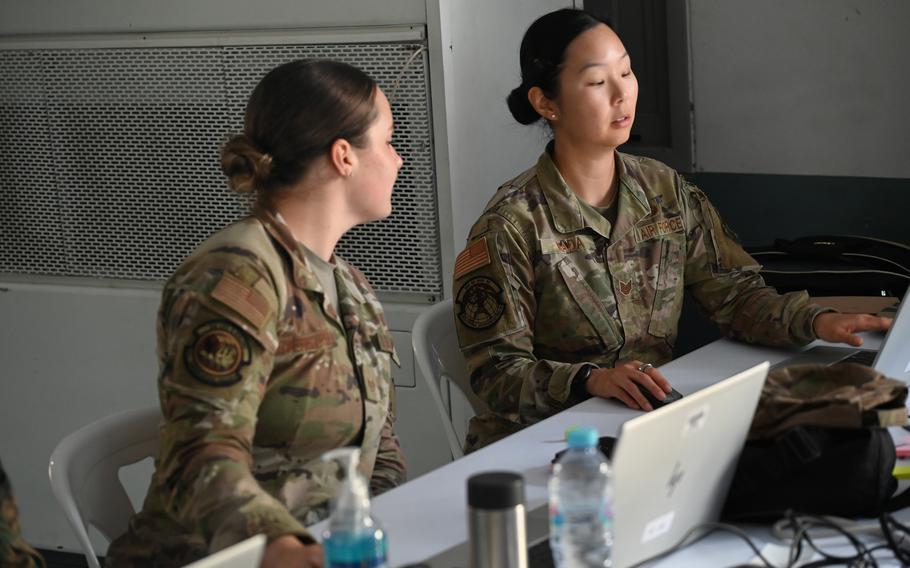 Two women in camouflage military uniforms sit at a table in front of laptop computers.