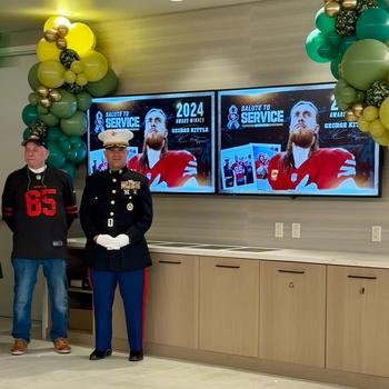 Two men stand before two TV screens that display a promotional image for Kittle’s award.