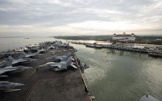 An aircraft carrier carrying aircraft sails in the water next to a dock.