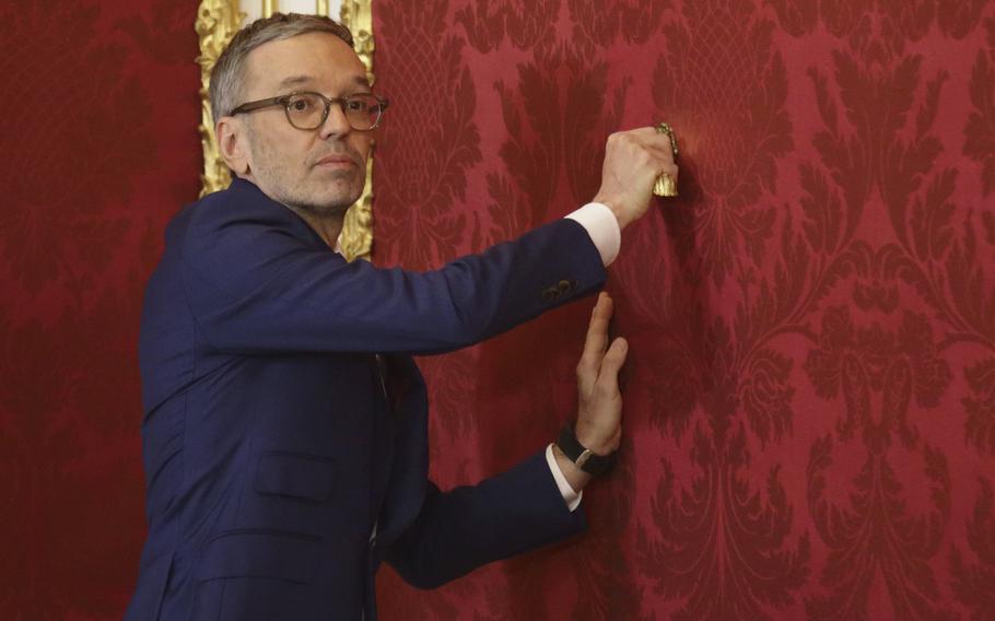 A man with glasses and a suit touches a red wall.