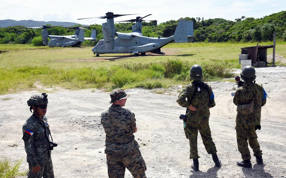 United States, Japanese and Filipino troops watch two Osprey land in a green field.