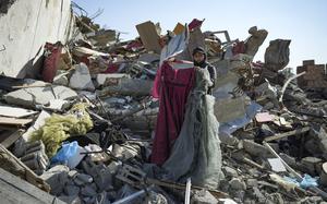 A woman holds two dresses while standing on a large pile of rubble.