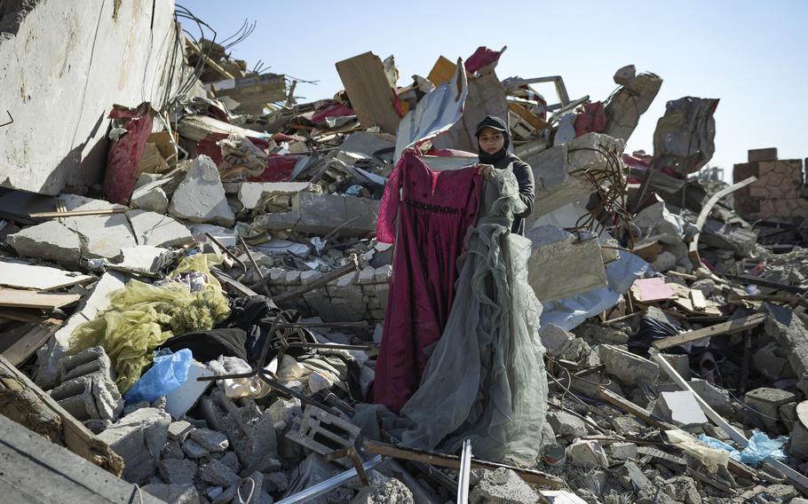 A woman holds two dresses while standing on a large pile of rubble.